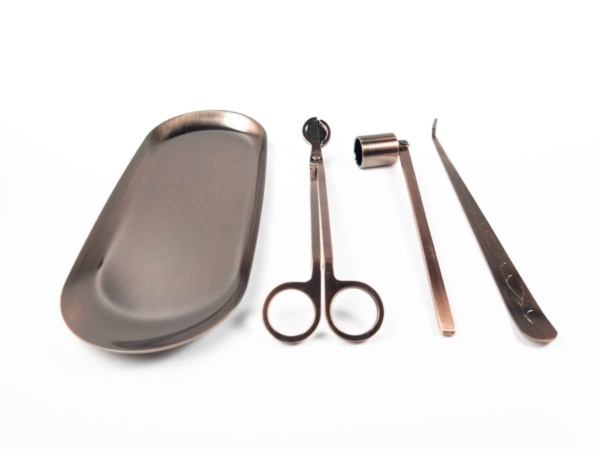 Brushed copper candle accessory set displayed on a white background. From left to right: a tray, wick trimmers, a snuffer, and a wick dipper, neatly arranged side by side.