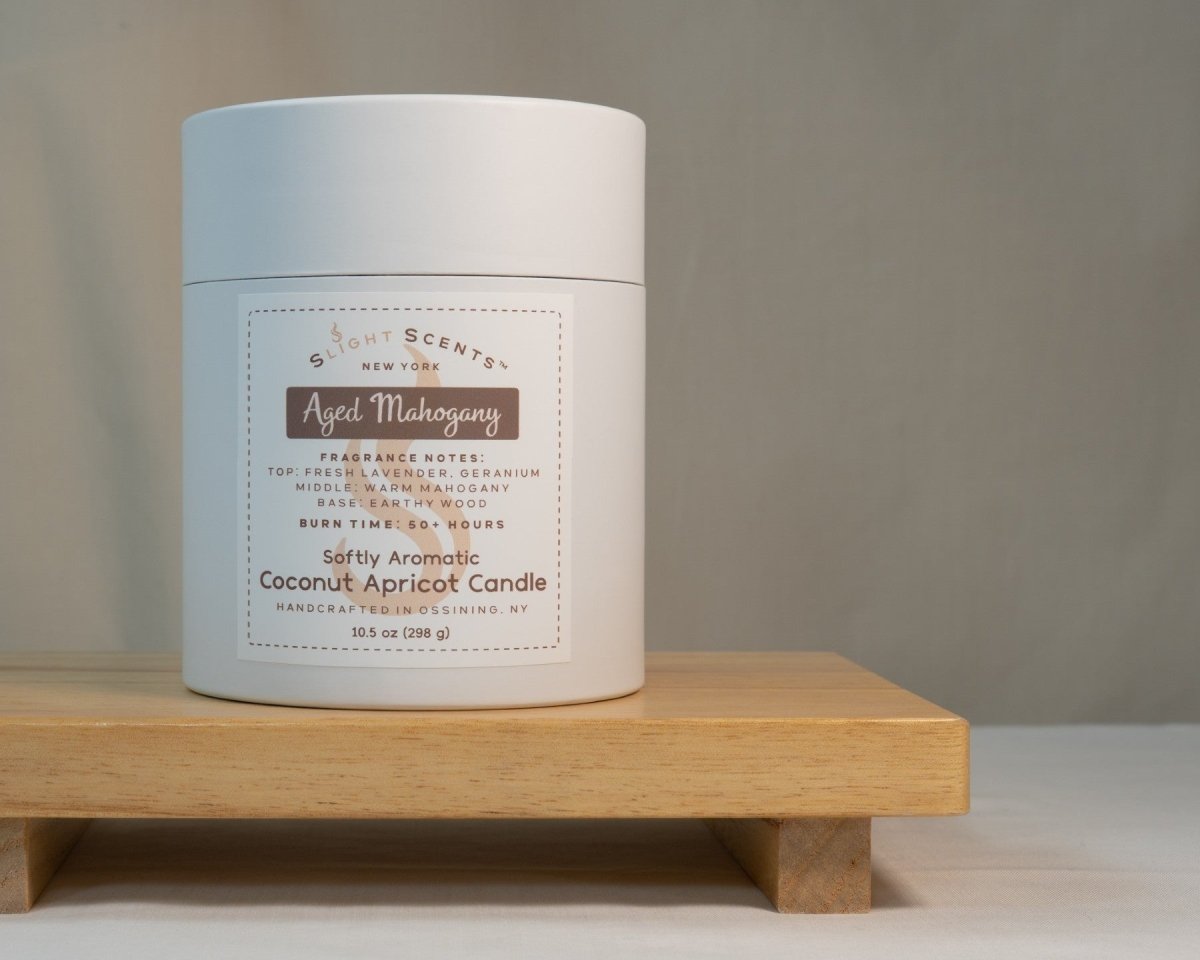White container for Slight Scents New York featuring the fragrance Aged Mahogany with notes of fresh lavender, geranium, warm mahogany, and earthy wood. Softly Aromatic coconut apricot candle, 10.5 oz, with a burn time of 50+ hours. Handcrafted in Ossining, NY.