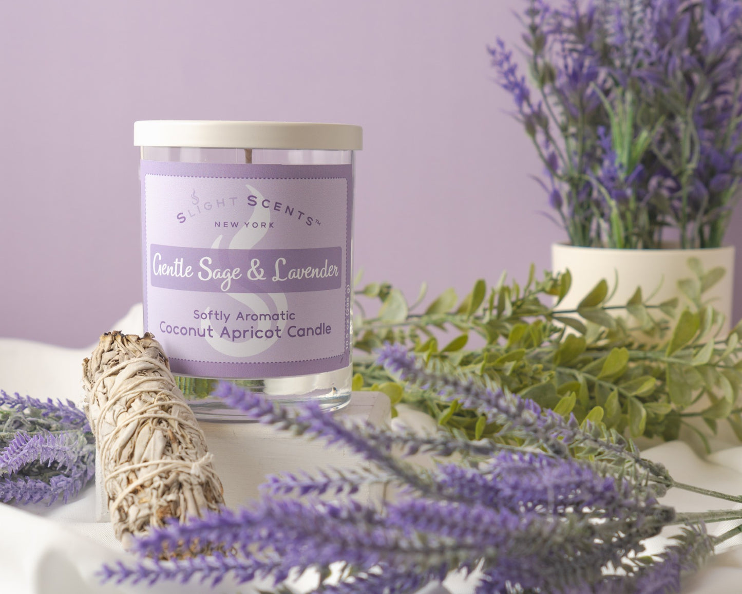 Gentle Sage & Lavender candle with a white sage smudge stick, lavender plants, and other green plants on a white surface.