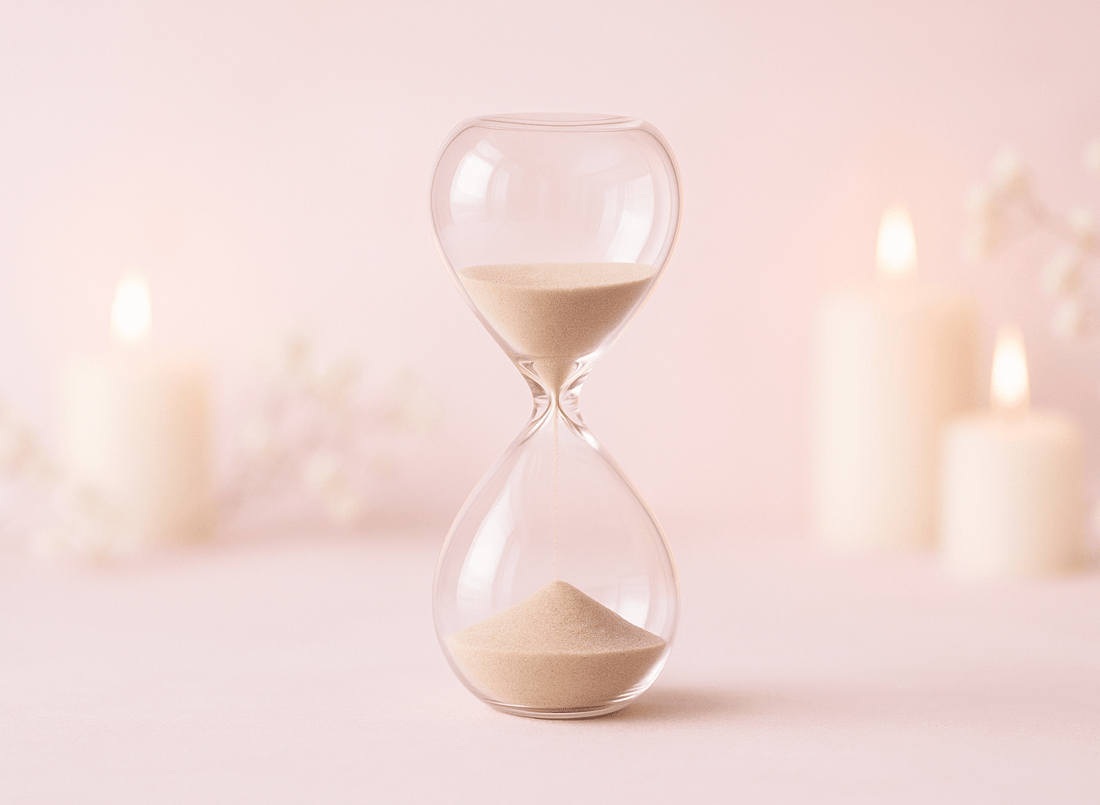 Glass hourglass with blurry pillar candles against a pink background.