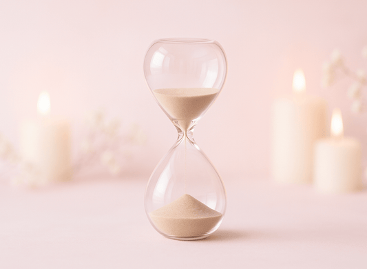 Glass hourglass with blurry pillar candles against a pink background.
