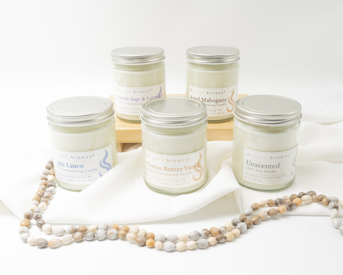 Five glass jar candles with silver lids and white labels are arranged on a white fabric backdrop with a small wooden riser. The labels show scents including Light Linen, Cocoa Butter Vanilla, Unscented, Gentle Sage & Lavender, and Aged Mahogany. 