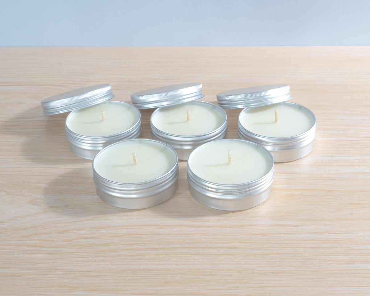 Five small silver metal tin candles with white wax are arranged on a light wood surface against a pale blue-gray background. Lightly scented soy discovery set. 
