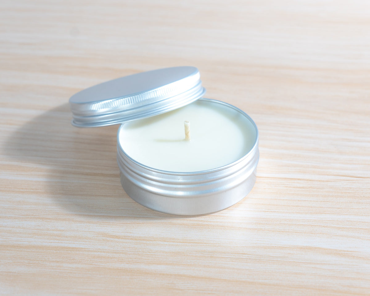 A small round silver metal tin candle with its lid off, showing a single wick and pale cream-colored wax, set on a light wood surface.
