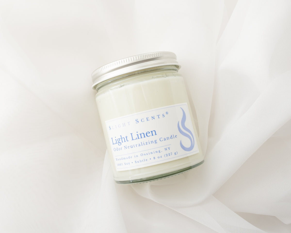 A glass jar candle with a silver metal lid and a white label reading 'Slight Scents Light Linen' on a soft white fabric background.