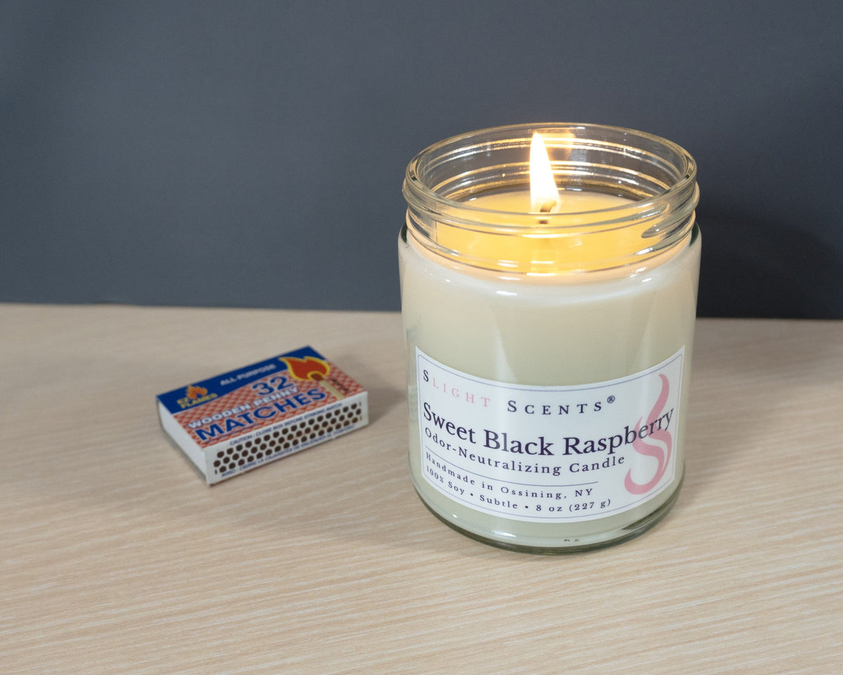 A lit Slight Scents soy candle labeled Sweet Black Raspberry, placed on a light wood surface next to a matchbox against a dark gray background.