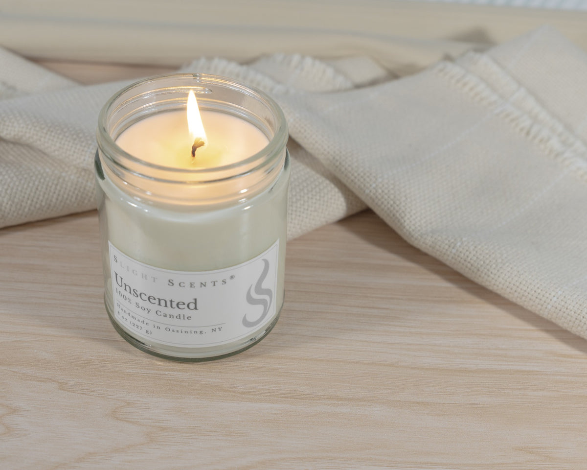 A lit Slight Scents Unscented soy candle in a clear glass jar, set on a light wood surface with a beige textured fabric in the background.