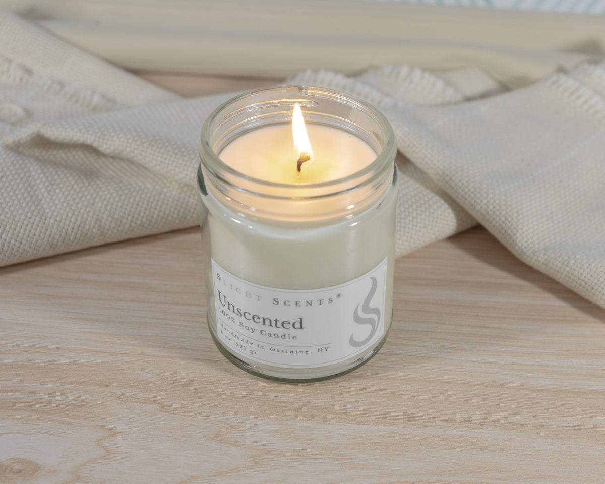 A lit Slight Scents Unscented soy candle in a clear glass jar, set on a light wood surface with a beige textured fabric in the background.