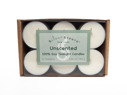 A 12-pack of 100% soy tealights neatly arranged in a cardboard box with a clear plastic cover, showcasing the tealights inside. The packaging displays a green label that says "Slight Scents New York Unscented 100% Soy Tealight Candles." 6.8oz 194g. Box turned on its side.